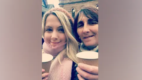 BBC/Gemma Sherlock A young girl with blonde hair and a hat, and a pink scarf, holds a cup, next to an older woman with reddish brown hair, also holding a cup, both are smiling into the camera 