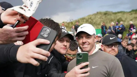 PA Rory McIlroy is smiling for a selfie with four or five young men. 

Rory is wearing a white nike hat and grey nike jumper. 

Behind him you can see the golf sand dunes and crowds of people gathering to see McIlroy. 

The sky is grey and the man closest to Rory is wearing a Yankee black cap and looking at his phone as he takes a selfie with Rory. 