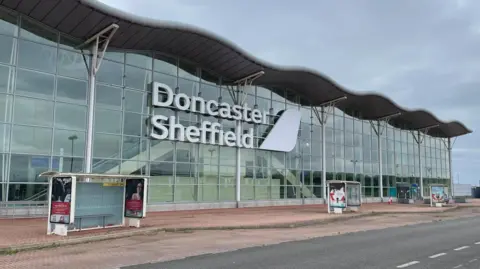 A large glass empty airport building with three bus stops outside.
