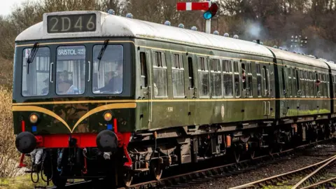 A steam locomotive that is dark green and has lots of windows. Steam is coming from funnels on top of it. A number above its cabin reads 2D46
