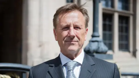Staffordshire County Council A man wearing a grey pin-striped suit, with a white shirt and a silver-coloured tie. He is standing outdoors against the backdrop of a stone building with black railings.