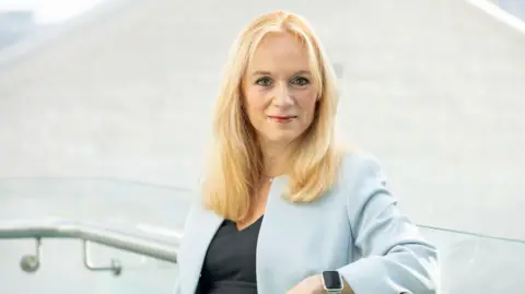 Grainge photography Victoria is looking straight at the camera. She has mid-length blonde hair. She is wearing a pale blue blazer, black top and smart watch. 