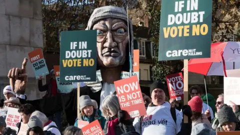 Getty Images Campaigners opposing assisted dying protest outside Parliament, in front of a big puppet of a judge. They hold signs holding slogans including "if in doubt vote no", "now is not the time" and "first do no harm".