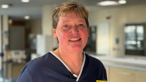 A woman with short light brown hair smiles at the camera. She is wearing a dark blue uniform with white trim in a V-neck. She has a yellow name badge with the NHS logo on it. She is stood in a room that has some cupboards, furniture and windows. 