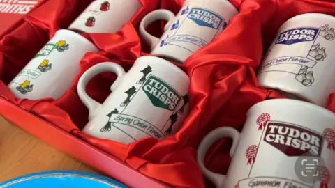 Six mugs with assorted  Tudor Crisps flavours embossed connected  the front, from "spring onion", successful  a greenish  font, to "salt and vinegar", successful  a bluish  font. All are displayed successful  a reddish  silk-lined box.