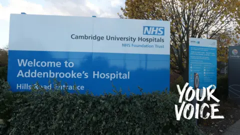 A sign at the entrance to Addenbrooke's Hospital. The top half is white with the logo of the Cambridge University Hospitals NHS Foundation Trust. The bottom half has the words "Welcome to Addenbrooke's Hospital" in white lettering on a blue background. There is a smaller blue and white sign to the right and a row of shrubs in front of the signs.