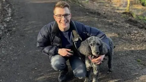 a young man wearing glasses wearing a parka jacket and jeans with his arm round a black dog. He is kneeling on a muddy path.