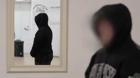 Alfie, whose identity is not being revealed, is wearing a black hooded top and is stood inside a room with his reflection visible in a mirror. HIs face is blurred and he has his hood up.