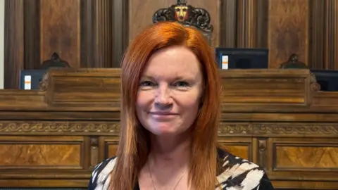 An image of a red-headed Ella Worthington, she has long hair and is smiling. She is standing in the council chamber, with wood panelling and seating behind her. She is wearing a black and white top and a necklace.
