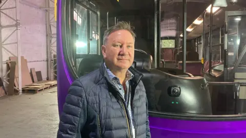 A man in a navy puffer jacket stands in a warehouse in front of a purple bus. Wood and cardboard are scattered in the background.