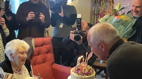 Avery Healthcare Group Joan is sat in a chair with Steve Bull and Steve Daley giving her a pretty iced cake topped in pink and purple flowers and candles. There are people looking on with cameras to capture the moment.