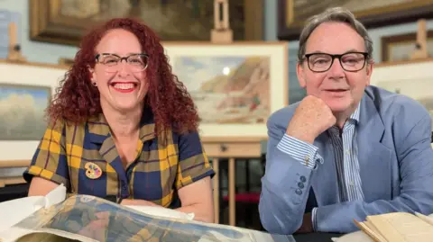 Samantha Harris and Eric Knowles are sitting in the museum with books and paintings in front of them. The background is blurred and there are pictures on the walls behind them and one painting is mounted on an easel.