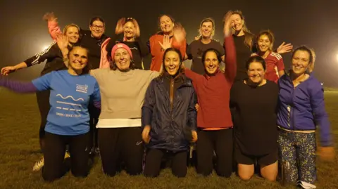 A group of 13 women at a football pitch smiling and waving at the camera.