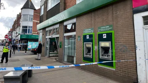 BBC The front of a high street branch of Lloyds Bank, which has had its front entrance smashed in by a car. Blue and white police tape surrounds the front of the bank. A police officer can be seen speaking on his radio behind the cordon. Another man in a black coat is standing in front of the damaged entrance.