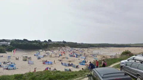 The image shows a wide sandy beach filled with groups of people sitting close to colourful windbreaks spread across the sand. Some people are walking along the shoreline while others are sitting or lying down on towels and beach chairs. There are a few small tents and beach shelters scattered around.