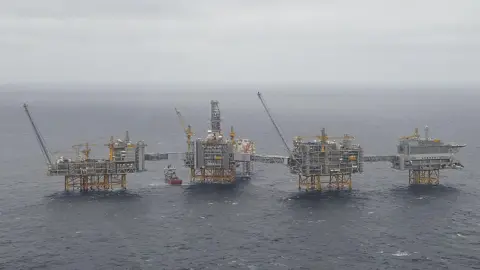 Reuters Four Equinor oil platforms in the North Sea. The grey platforms have yellow legs and cranes and a red and white boat is sailing between two of them. 