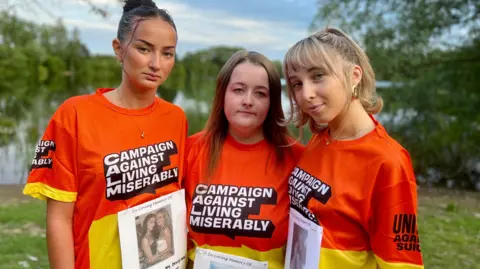 Nikki Fox/BBC Alicia Down, Charlotte Millar and Emily Ferguson wearing orange T-shirts with Campaign Against Living Miserably across the front and a photo of Amelia pinned on their front. They have their arms around each other and are standing in a green space. 