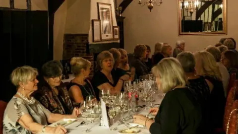 The Strangers Club About 24 women seated either side of a long table laid out for a formal dinner. The table has a white tablecloth and is covered with wine glasses. They are in a dimly lit room with a mirror on the wall at the opposite end of the image, and a portrait of the late Queen hanging above an old fireplace.