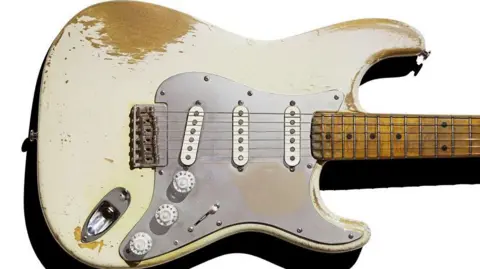 PA Picture of a white electric guitar looking well used with three dials and the paint peeling off revealing the wooden base beneath