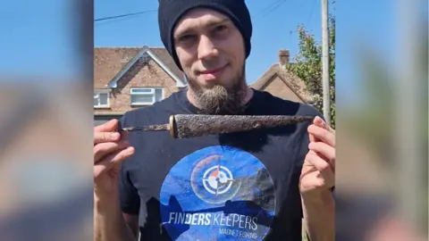 Grant Osborne Grant Osborne looks at the camera. He wears a black beany hat and a tee-shirt reading Finders Keepers Magnet Fishing with a compass logo. He holds a rusted triangular dagger.