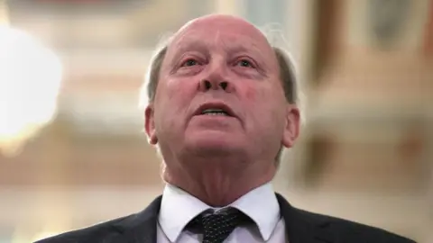 PA Media A head and shoulders shot of Jim Allister MP show him with his chin tilted upward, nostrils flared and his eyes red. He is wearing a black suit jacket, black tie with white spots and a white shirt. The top of his head is bald but his hair is grown out on on the sides of his head.