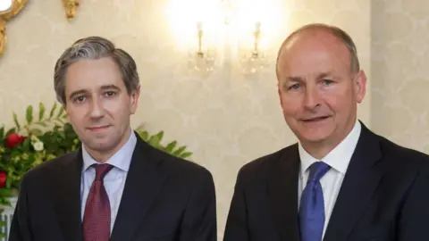 PA Media Simon Harris and Micheál Martin. Simon has grey-ish hair and wearing a black blazer, light blue shirt and burgundy tie. Micheál is balding and wearing a black blazer, white shirt and blue tie. Both are looking at the camera and a floral arrangement can be seen in the background. 