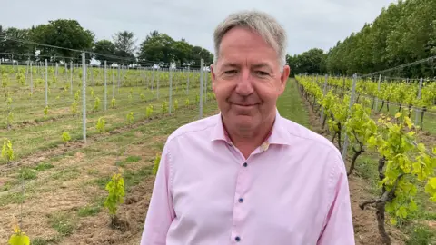 Man with short grey hair smiling at the camera. He is wearing a pink button down shirt and standing in front of his vineyard.