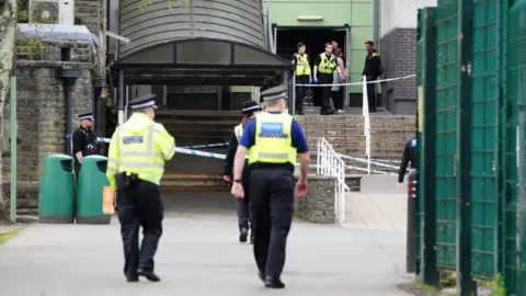 Two police officers are seen with their backs to the camera outside Ysgol Dyffryn Aman school. There is a police cordon in the background with more police gathered at the top of a set of stairs.