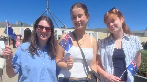 Three young women holding small Australia flags and smiling