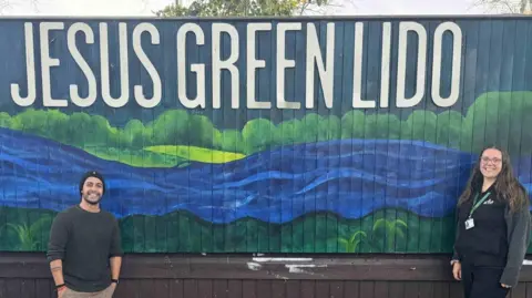 GLL Anugrah Mishra and Annabel Wright, both smiling broadly, standing against a fence which has been painted with a blue and green mural showing an undulating bright blue river with trees and fields above and grass below. It says Jesus Green Lido in capital letters above. Anugrah Mishra is on the left, has a close cropped dark beard and is wearing a black beanie hat and grey jumper and his hands are in his pockets. Annabel Wright is on the right and has long dark brown hair over a black fleece and is wearing glasses. 