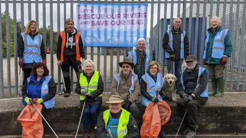 Grantham RiverCare A group of litter pickers wearing blue, yellow and orange high vis vests, with some standing on top of a wall and others sitting below it. They are holding litter picks and bags and there is also a golden retriever. A sign on the fence says 'let's rescue our rivers and save our seas'.