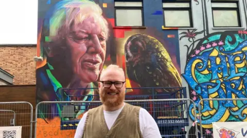 Guthrie O'Brien A man with a reddish beard, glasses and wearing a white T-shirt and beige waistcoat - is pictured standing in front of a large mural on the side of a building. The mural is of Sir David Attenborough looking - slightly askew - at a tawny owl. 