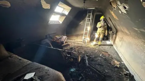 ECFRS A firefighter dressed in yellow with breathing apparatus stands in a bedroom which has been left totally blacked by fire. The carpet is burned, the walls are thick with soot and the floor is littered with burned cushions, clothes and furniture.