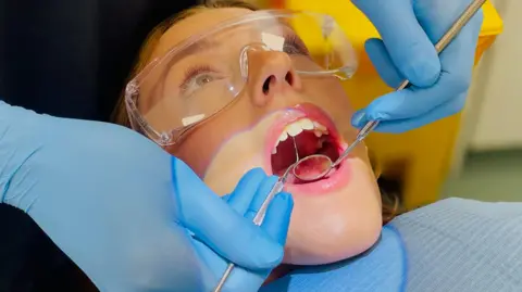A woman lies back in a dentist chair with her mouth open. She is wearing protective glasses with the dentist examining her teeth with a mirror and dental probe 
