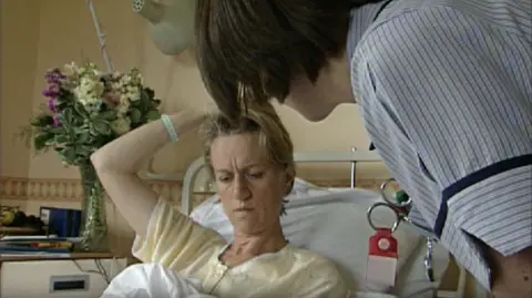 Sally Taylor in hospital in 1999. She is in a hospital bed being tended to by a nurse.