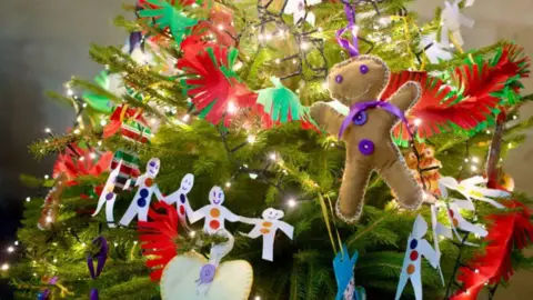 A Christmas tree decorated with a variety of festive decorations, including bright fairy lights, tinsel and a stitched gingerbread man figure.