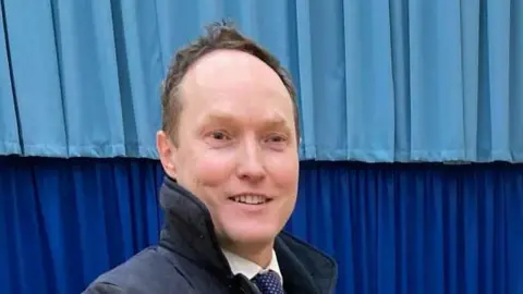 Robin Hunter Clarke Scott Hussey is wearing a dark blue coat over a white shirt, blue tie with white spots. He has brown hair and is smiling while looking off camera 