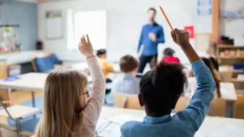 Getty Images Two girls in classroom with raised hands, other pupils and a male teacher are visible in the background