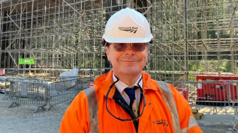 The picture shows a man in a bright orange hi-vis coat and white protective safety helmet both with Network Rail branding on the front. There is a lot of building and construction work going on behind him with lots of scaffolding. There is also a generator behind him to the right hand side.