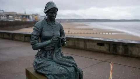 The bronze sculpture is of a woman sitting on the plinth of the sculpture knitting. The bronze has weathered to a bluey black colour. The beach at Bridlington can be seen in the background.