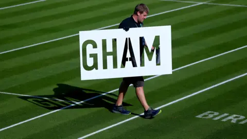 Edgbaston Priory A groundsman dressed in black polo shirt and shorts is holding a large stencil showing GHAM as he walks off a grass tennis court