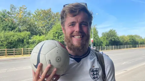A man wearing a white England football shirt is standing by the side of a road holding a white Nike football.