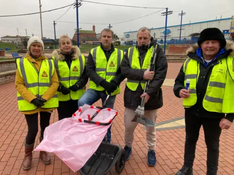 Two women and three men wearing hi-vis yellow jackets standing in a row in on a seaside promenade with a trolley topped with a big pink plastic bag. The promenade is tiled with red bricks and a large warehouse-like building can be seen in the background.