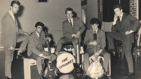 Five young men photograhped in the 1950s wearing suits typical of that period. Two are holding guitars, one is standing over a drumkit with "The Cyclones" written on the front.
