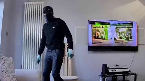 Essex Police A man dressed in black with a balaclava standing in a living room with the TV on behind him.