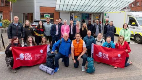 Birmingham Children’s Hospital Charity More than 20 people posing for the camera outside a building. Mike Weaver (centre right) has one knee on the ground and is wearing an orange top. Two identical red and white banners, including the words Kili for kids, are being held by other people. 