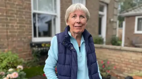 A woman wearing a blue gilet is looking into the camera and standing in front of the window of a house. She has short grey hair and is wearing a turquoise quarter-zip long-sleeve top.