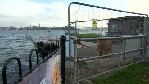 BBC On the left is a black fence leading to a silver gate blocking access to the Mount Edgecumbe slipway. At the centre the slipway is light brown and there are red cones lining it. On the right is a building.