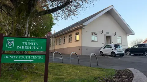 Exterior view of Trinity Parish Hall, a single‑storey cream building with parked cars, beside a green sign reading “Welcome to Trinity Parish Hall” and “Trinity Youth Centre,” under a large tree.