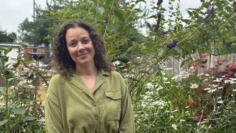 A woman with long brown hair and a green top stood in a garden